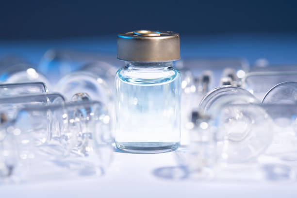 There is a full vaccine tube in the middle of the white background. There are a few empty vaccine tubes around it. Blue light on the background.