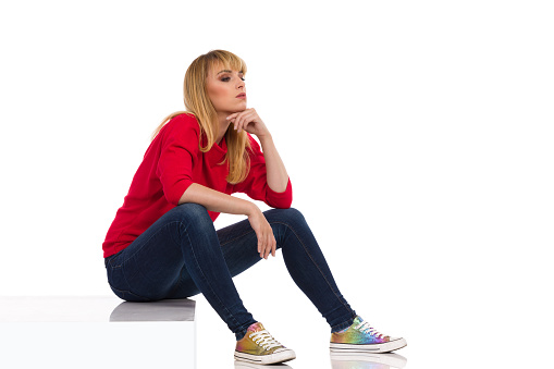 Bored young woman in red sweater is sitting on a step, holding hand on chin and looking away. Side view. Full length studio shot isolated on white.