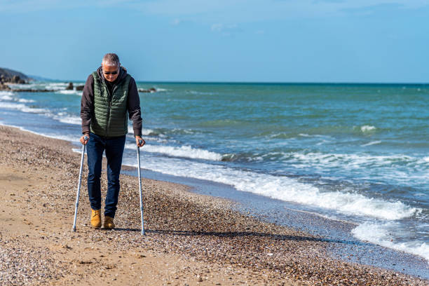 Disabled adult man using aluminum forearm crutches by the sea Stylishly dressed disabled adult man using aluminum forearm crutches as a mobility aid to walk on the seaside in Turkey. Full length view. aids walk stock pictures, royalty-free photos & images