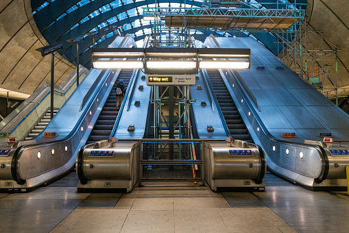 Color image depicting a single commuter on an elevator amid the glass and steel design of the architecture at Canary Wharf underground station in London. An escalator leads up to the skyscrapers and towers of the business district beyond.