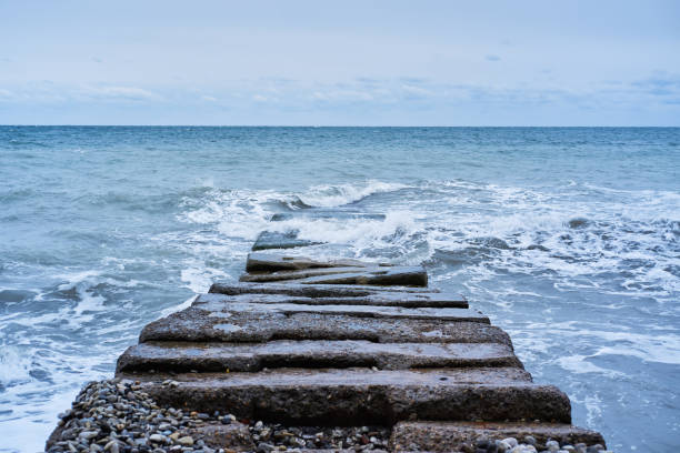 Long concrete breakwater stretching away into depths of stormy blue sea. Waves break on rocks and fly apart into small drops and turn into foam. Banner of sea landscape.