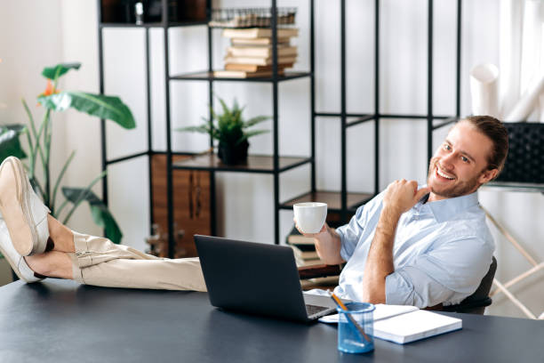 istock Smiling attractive young man, freelancer or manager, is relaxing at workplace with legs thrown on table. Modern guy uses laptop, browses the Internet, looks for ideas for a project, while sitting at home office 1306674904