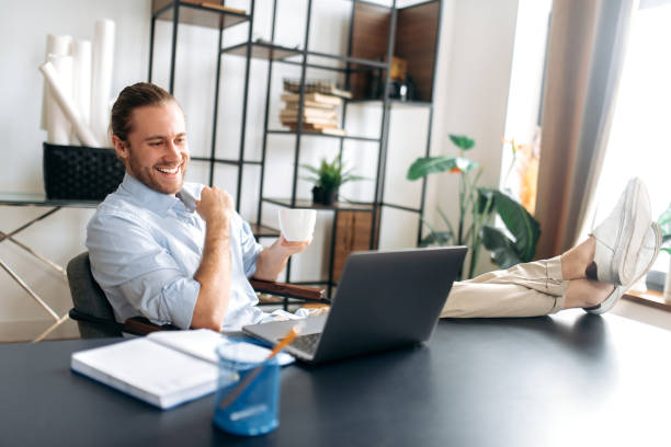 istock Smiling attractive young man, freelancer or manager, is relaxing at workplace with legs thrown on table. Modern guy uses laptop, browses the Internet, looks for ideas for a project, while sitting at home office 1306674911