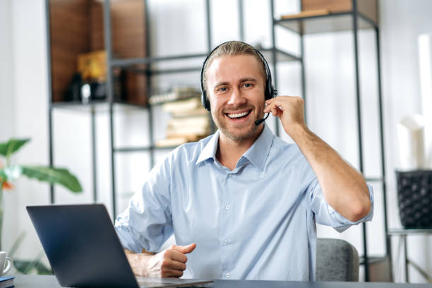istock A friendly confident caucasian guy, employee, freelancer or call center operator, wearing a headset, sits at his workplace in the office and looks at the camera with a smile 1306674926