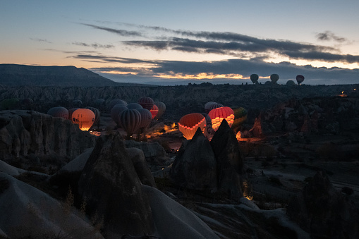 Hot air balloons glowing in early morning while inflating among rocks of mountain landscape in Cappadocia. Colorful illuminated balloons in blue hour of sunrise. Travel Goreme Turkey, 01 09 2021