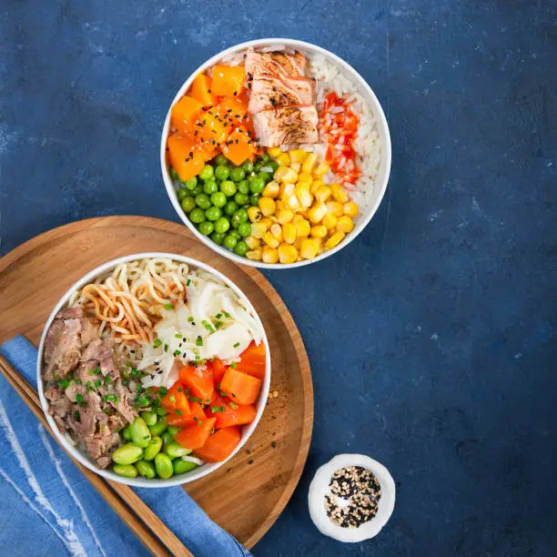 Two assorted poke bowls, flamed salmon, pulled pork, vegetables, rice, sauces. Top view, close-up. Hawaiian dish, blue dark background. Healthy and clean eating concept. Trendy asian food.