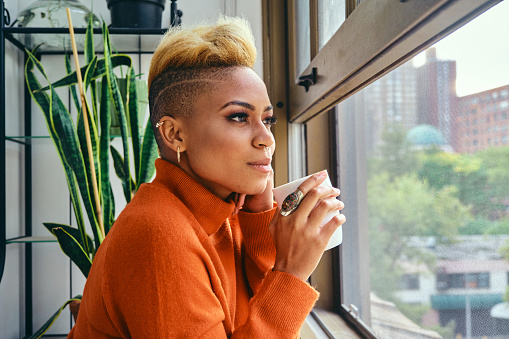 Closeup of thoughtful young female hipster looking through window while having coffee at home