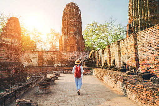 Young woman tourist in a straw hat and white clothes walking with light city backpack through Ayutthaya Wat Phra Ram ancient ruins streets in Thailand. History, tourism, sightseeing concept.