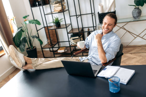 istock Happy caucasian young man, freelancer or manager, is relaxing at workplace with legs thrown on table. Stylish guy uses laptop, browses the Internet, looks for ideas for a project, while sitting at home office 1306691426