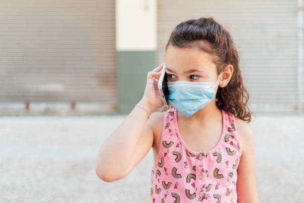 a girl in a tank top and facemask communicates with her family, friends and grandparents outdoors in pandemic summer with her cell phone. concept of long-distance communication. - tank top fotos imagens e fotografias de stock