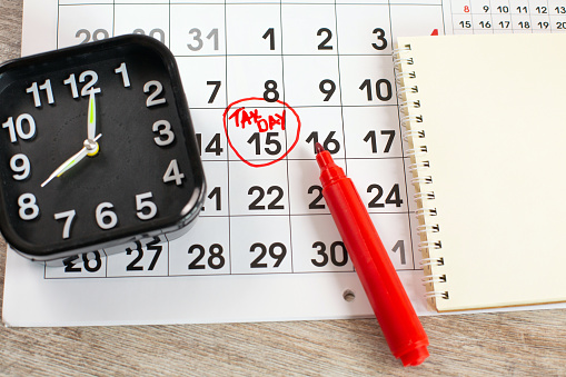 Writing Tax day on 15th April monthly calendar circled in the circle in a red marker with notebook and alarm .