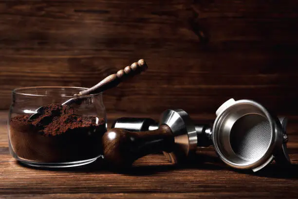 Photo of glass jar with ground coffee temper portofilter wooden coffee spoon on natural wooden brown background close-up copy space