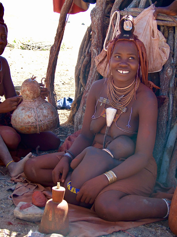 Closeup photo of a smiling Himba woman nursing her young child on her lap, sitting on the ground outside her hut in northern Namibia. The woman is wearing traditional skirt, jewelery, hairstyle and head dress denoting her married status. On the left another woman, partially seen, moves a gourd filled with cow's milk back and forth to make butter.