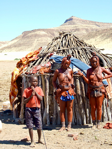 Vertical photo of three Himba people standing outside their family hut in a small village in northern Namibia. The women are dressed in traditional clothing, wearing jewelery and have clay covered hairstyles and a cowhide headpiece denoting their married status. In the background the arid hills of this dry desert landscape.