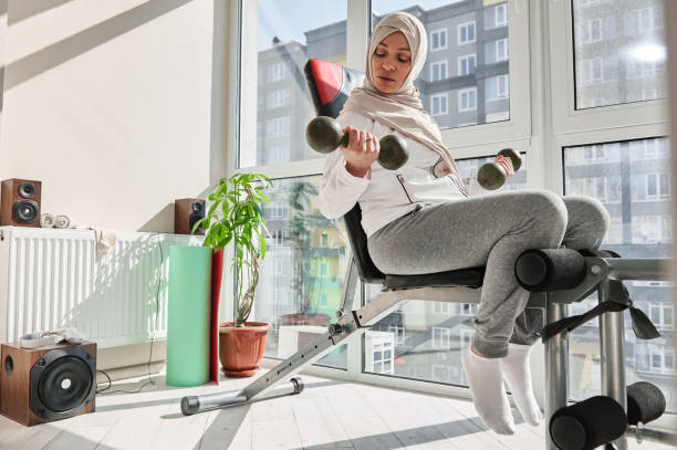 giovane donna attraente in hijab, esegue esercizi con manubri, seduta su una panchina in palestra di casa. - women weight bench exercising weightlifting foto e immagini stock