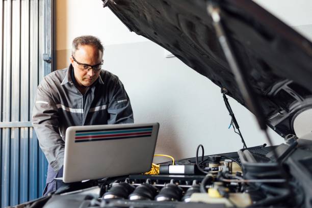 istock Car mechanic in his workshop diagnosing a broken-down car with his laptop. Person fixing a broken car. Shop clerk with safety glasses. 1306707727
