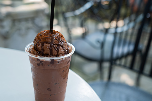a glass of iced dark chocolate, one topped with chocolate ice cream, glass placed on white table with cafe environment.