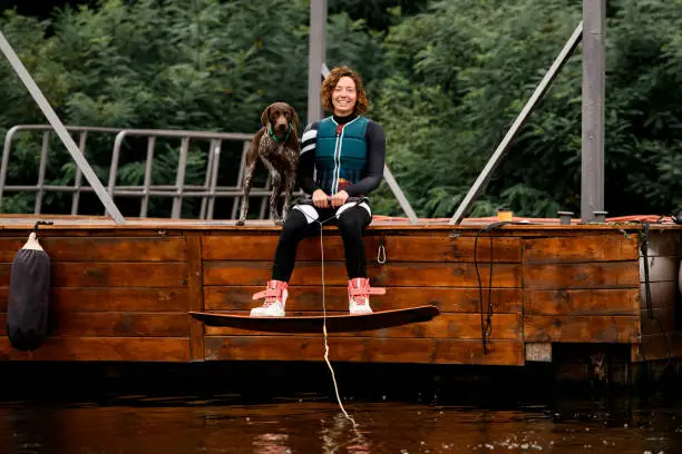 young smiling woman sits on wooden pier with wakeboarding board on her legs and rope in her hands. Dog standing nearby