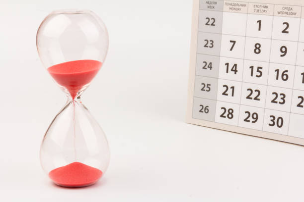 istock Time passing concept. Crystal hourglass with red sand and calendar on light background 1306714799