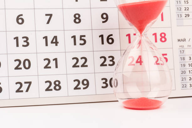 istock Time passing concept. Crystal hourglass with red sand and calendar on light background 1306714835