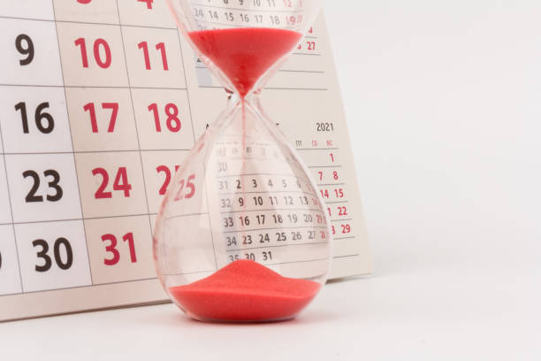 istock Time passing concept. Crystal hourglass with red sand and calendar on light background 1306714848