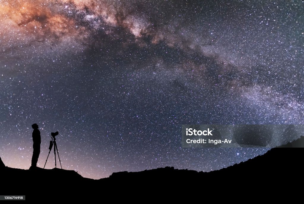 The man silhouette with camera and tripod, stands on the hill and looking at the bright milky way galaxy.  Beautiful night landscape. Astronomy Stock Photo
