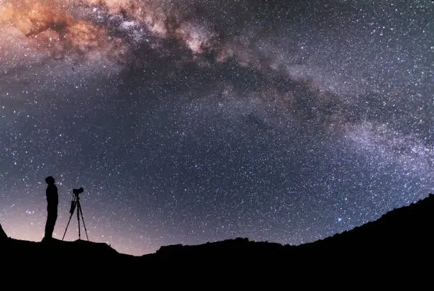 Photo of The man silhouette with camera and tripod, stands on the hill and looking at the bright milky way galaxy.  Beautiful night landscape.