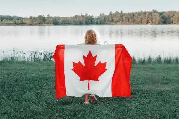 Girl wrapped in large Canadian flag by Muskoka lake in nature. Canada Day celebration outdoor. Kid in large Canadian flag celebrating national Canada Day on 1 of July.