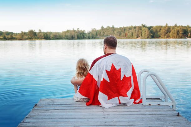 vater und tochter in große kanadische flagge gewickelt sitzen auf holzsteg am see. canada day feier im freien. vater und kind sitzen am 1. juli zusammen, um den nationalen kanadatag zu feiern. - canadian flag fotos stock-fotos und bilder