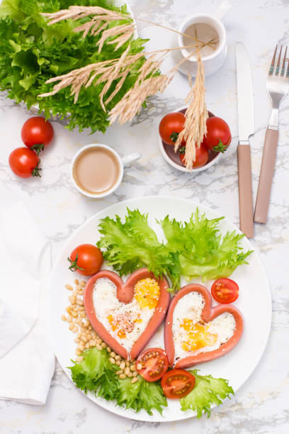 istock Romantic breakfast. Fried eggs in heart shaped sausages, lettuce and cherry tomatoes on a plate on the table. Top and vertical view 1306723881