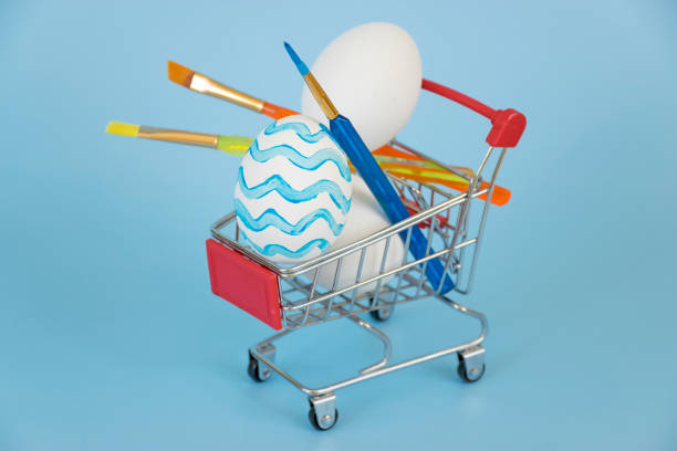 Easter egg decorated in blue waves with other white eggs and colorful paintbrushes in shopping cart on blue background. Preparation and online shopping for Happy Easter holiday.