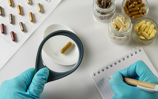Hand of a pharmacist holds a capsule with dietary supplements and magnifying glass on the background of jars of ingredients in laboratory