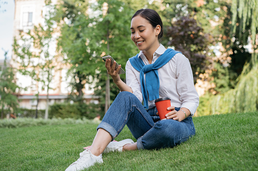 Beautiful asian woman using mobile phone, communication online, holding cup of coffee, sitting on grass. Young smiling student studying, reading, e-learning