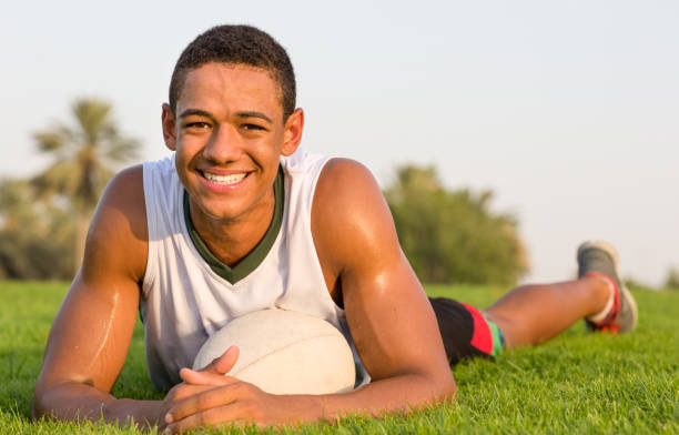 Happy fit black teen athlete lying on the grass with a rugby ball and smiling to the camera. Portrait of smiley African young man relaxing on the grass with a rugby ball at the park. the black ball stock pictures, royalty-free photos & images