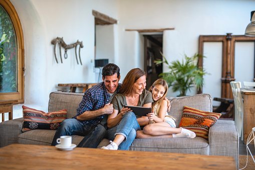 Front view of laughing Spanish father trying to put arm around wife and daughter on sofa in family home while using digital tablet.