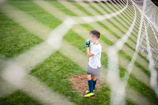 Personal perspective through netting of Caucasian 8 year old male goalie waiting and watching action on field.