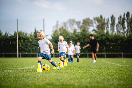 Low angle view of mature male coach directing boys and girls aged 5-9 years as they do dribbling drill around pylons during sports training camp.