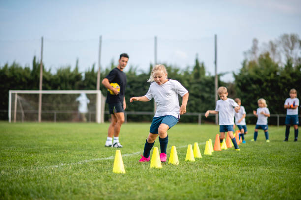 Coach Watching Action of Girl Footballer Doing Agility Drill Focused 7 year old girl moving around row of pylons as she and teammates do agility drill under direction of male coach during sports training camp. 6 11 months stock pictures, royalty-free photos & images