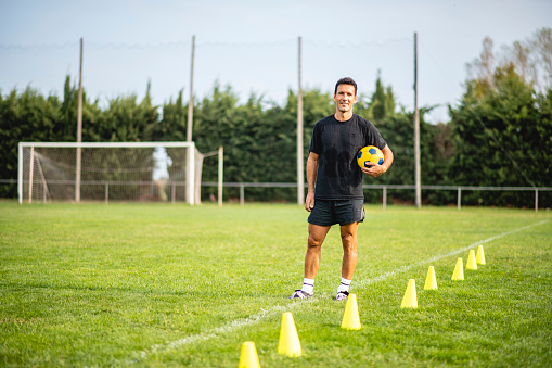 Outdoor portrait of mature male coach smiling at camera while standing on field with ball under arm in preparation for sports training drill.