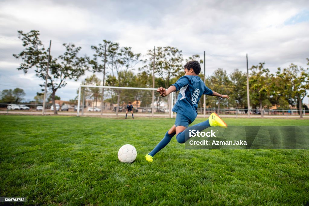 Athletic Mixed Race Boy Footballer Approaching Ball for Kick Rear view of agile 13 year old boy athlete wearing blue uniform and approaching ball for kick toward goal. Soccer Stock Photo