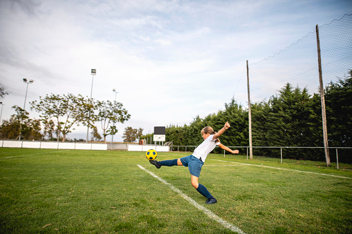 Young Caucasian female athlete wearing white sports shirt, blue shorts, and blue shoes kicking ball on field with arms outstretched.