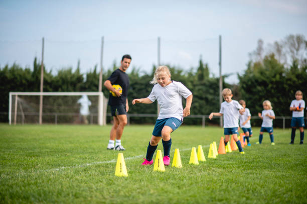 Coach Watching Action of Girl Footballer Doing Agility Drill Focused 7 year old girl moving around row of pylons as she and teammates do agility drill under direction of male coach during sports training camp. drill team stock pictures, royalty-free photos & images