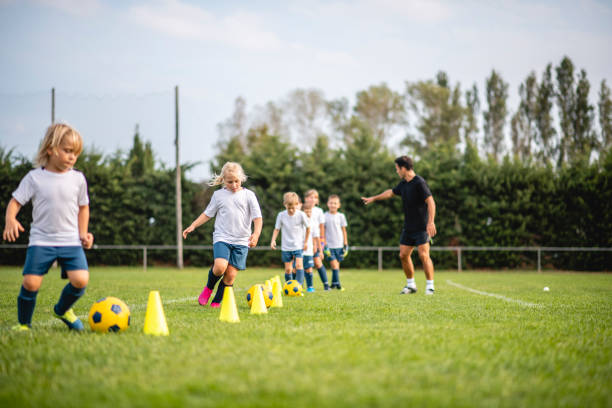 Pre-Adolescent Footballers Dribbling Around Pylons Low angle view of mature male coach directing boys and girls aged 5-9 years as they do dribbling drill around pylons during sports training camp. 6 11 months stock pictures, royalty-free photos & images
