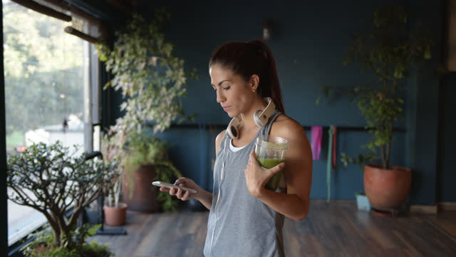 Wanita muda Amerika Latin minum jus hijau di gym sambil memeriksa pesan di smartphone dengan headphone di lehernya - konsep gaya hidup sehat