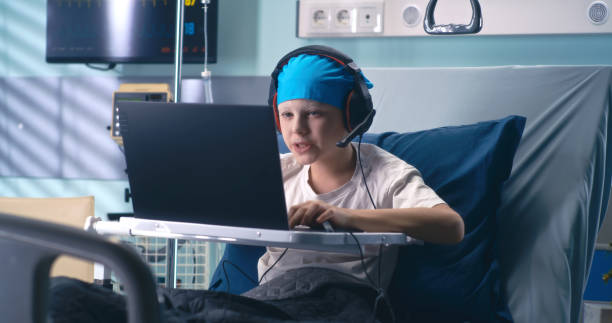 Gamer in headset playing in hospital ward Concentrated boy with headset using laptop while playing videogame on laptop in ward of oncology clinic real time stock pictures, royalty-free photos & images