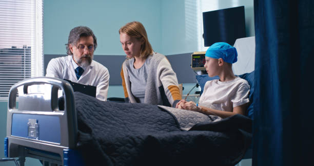 Middle aged oncologist explaining treatment prospects to woman and boy Mature bearded man discussing good therapy prospects with mother of cancer patient in ward on contemporary oncology clinic real time stock pictures, royalty-free photos & images