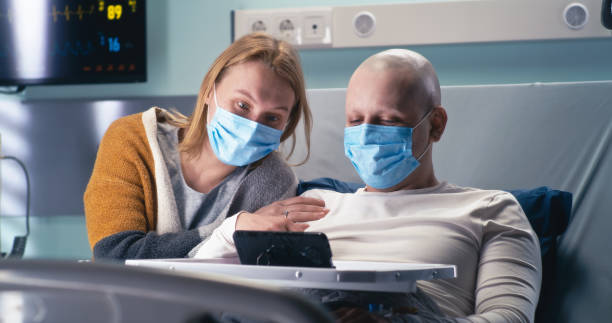 Man and woman in masks making video call from clinic Bald cancer patient in mask and wife speaking with relatives on laptop in modern ward of oncology hospital during pandemic real time stock pictures, royalty-free photos & images