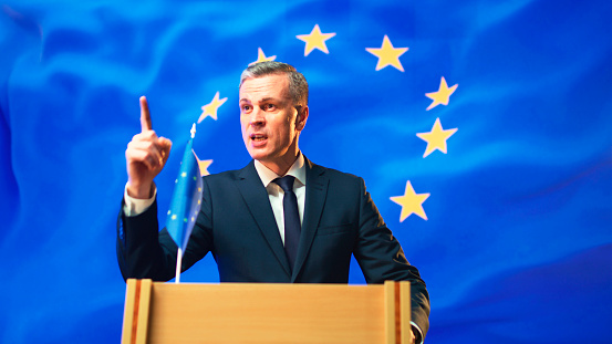 Confident mature man in suit gesticulating and asking questions to audience while standing on rostrum against flag of Europe