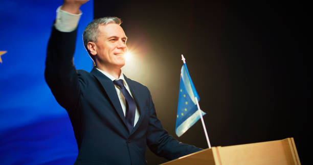 Positive mature man in suit waving hand and smiling to audience while standing on rostrum against European flag against photo camera flashes
