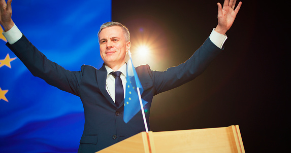 Friendly middle aged man in suit waving hand and clasping hands over head while greeting audience from stage against European flag